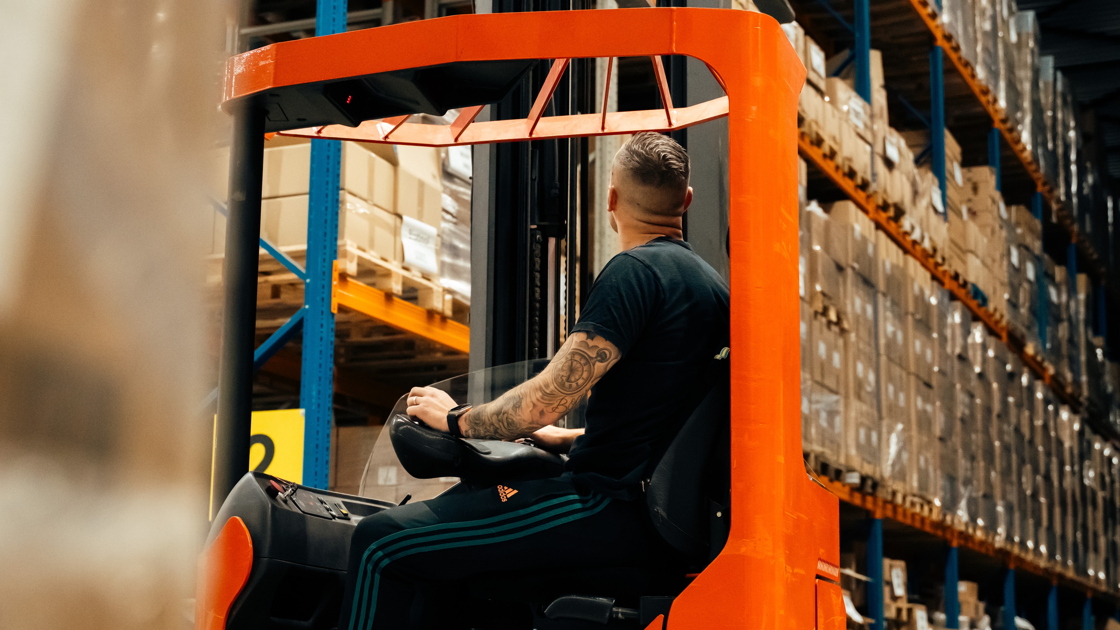 Medewerker met tatoeages rijdt op een oranje reachtruck tussen de stellingen