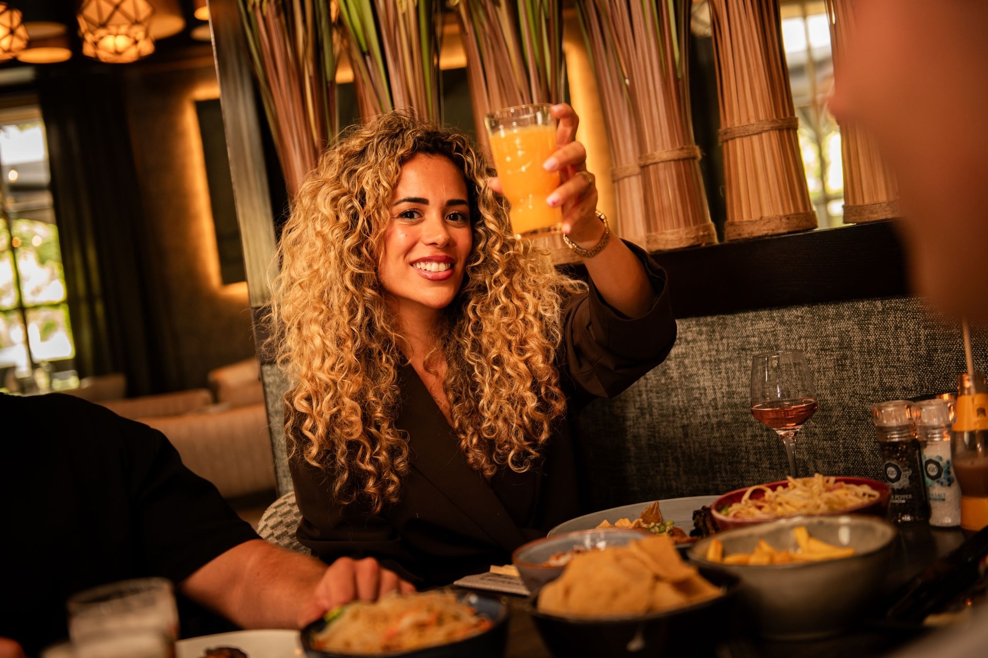 Vrouw heft lachend een glas sinaasappelsap aan tafel bij Grand Plaza